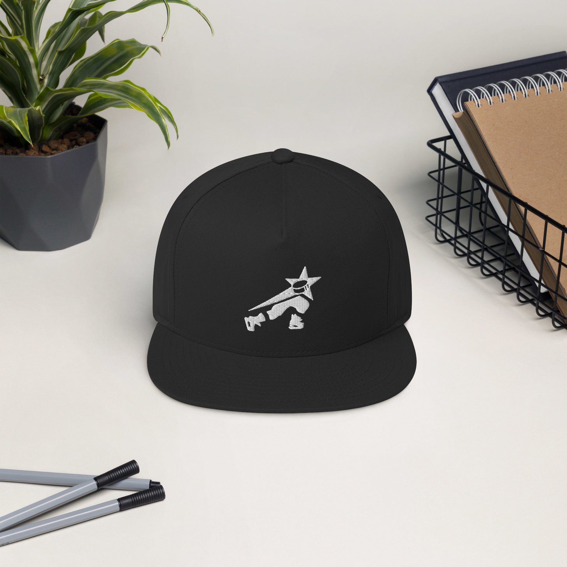 Black snapback cap with embroidered white hockey player design on a desk with pencils, notebook, and a potted plant.